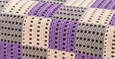 A bed-sized crochet blanket made of purple, gray, and cream block squares in a geometric patchwork design, draped over a bed.
