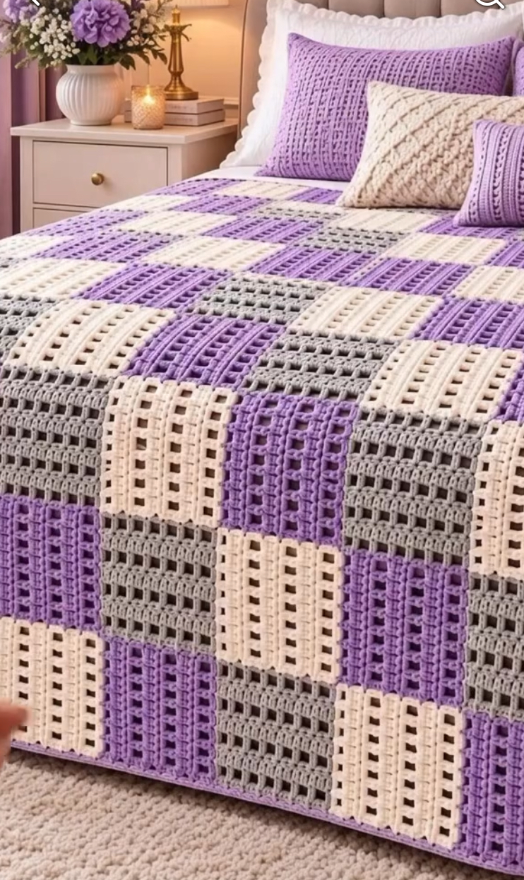 A bed-sized crochet blanket made of purple, gray, and cream block squares in a geometric patchwork design, draped over a bed.