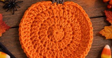 A crochet pumpkin coaster with a round orange body and brown stem, styled with pumpkins, leaves, and Halloween decorations.