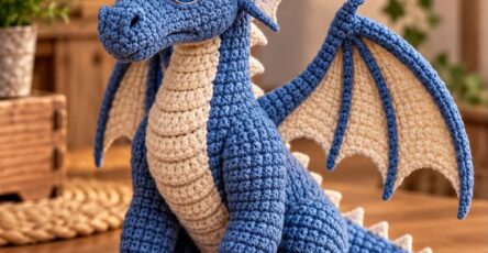 A crocheted blue and cream dragon amigurumi with detailed wings, horns, claws, and expressive eyes sitting on a wooden table.