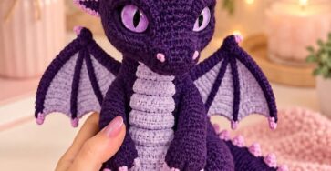 A crocheted purple dragon amigurumi with lavender belly, pink spikes, and detailed purple wings, posed among floral decor and soft lighting.