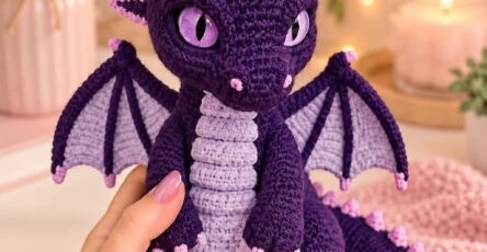 A crocheted purple dragon amigurumi with lavender belly, pink spikes, and detailed purple wings, posed among floral decor and soft lighting.