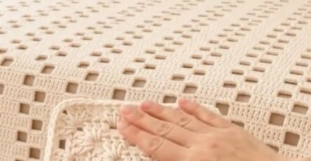 A hand holding a cream-colored crochet granny square in front of a matching crochet bedspread with openwork square motifs on a neatly made bed.
