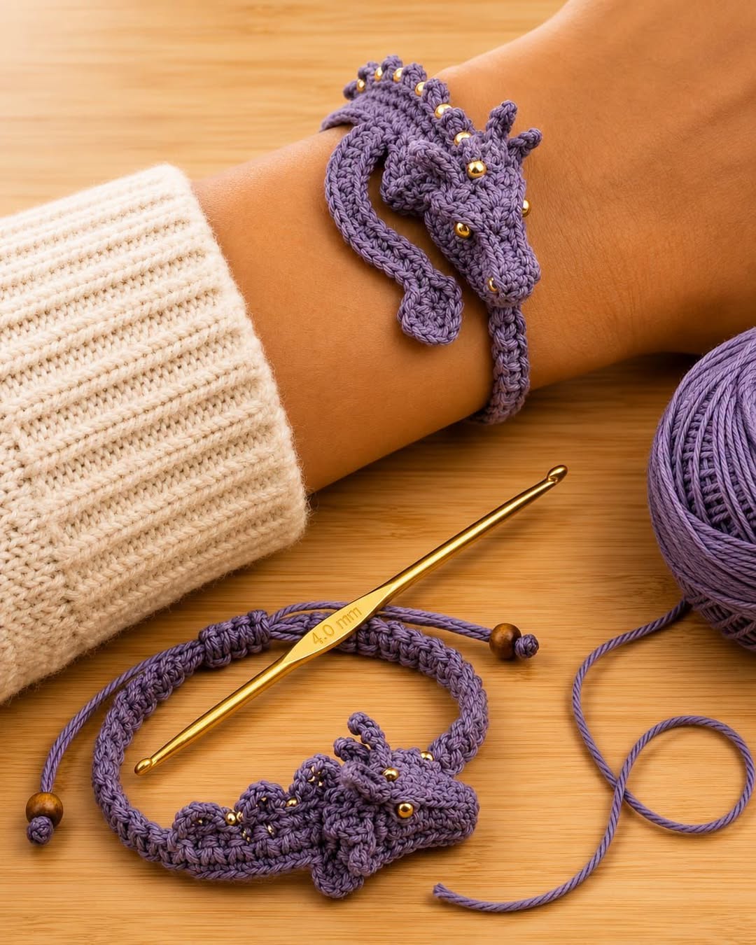 A lavender crochet bracelet shaped like a dragon with gold bead details, shown worn on a wrist and laid flat beside a crochet hook and yarn.