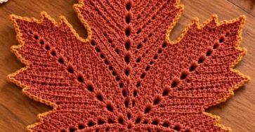 A crocheted maple leaf in autumn colors on a wooden table, surrounded by fall decorations.