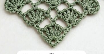 A green triangular crochet motif with shell stitches and a lacy, openwork pattern, with a ball of yarn, crochet hook, and a plant nearby.