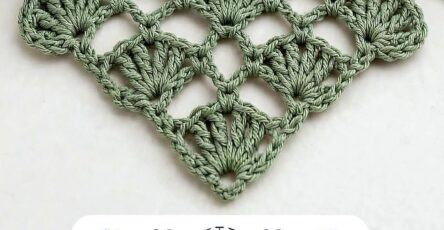 A green triangular crochet motif with shell stitches and a lacy, openwork pattern, with a ball of yarn, crochet hook, and a plant nearby.