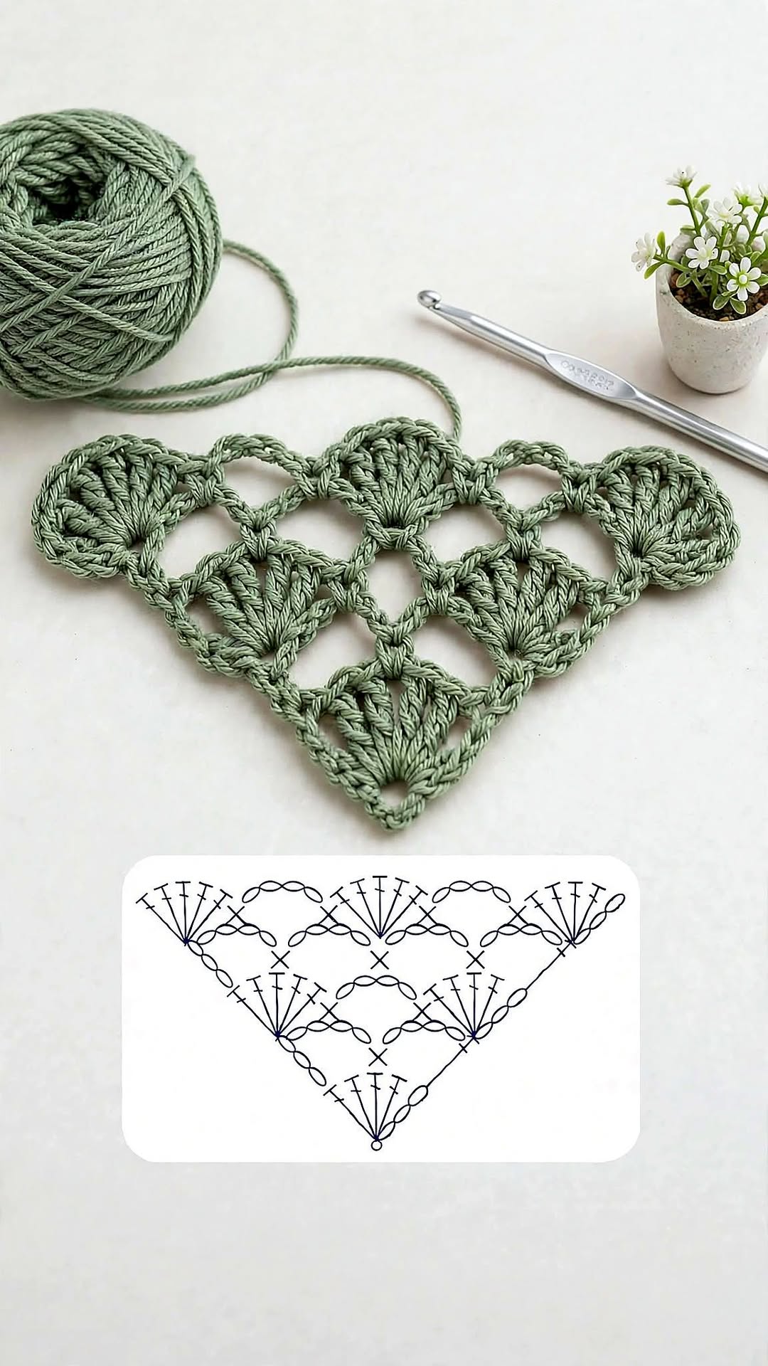 A green triangular crochet motif with shell stitches and a lacy, openwork pattern, with a ball of yarn, crochet hook, and a plant nearby.