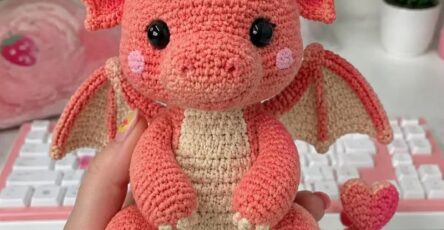A hand holding a crocheted amigurumi dragon plushie with coral and cream colors, wings, horns, and heart-shaped details.