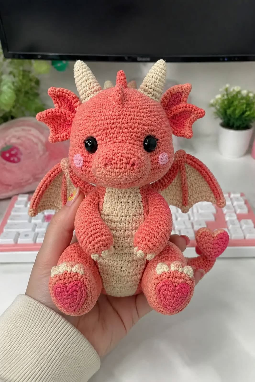 A hand holding a crocheted amigurumi dragon plushie with coral and cream colors, wings, horns, and heart-shaped details.