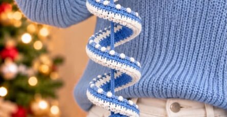 Hand holding a crocheted blue and white spiral Christmas ornament with Santa faces, holly leaves, and a tassel, in front of a Christmas tree.