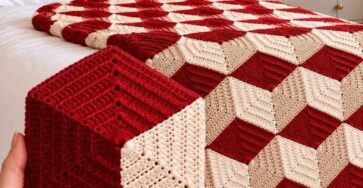 A geometric crochet blanket and matching cube pillow in red and cream, creating a 3D cube illusion, displayed on a bed with tassels on the blanket edge.