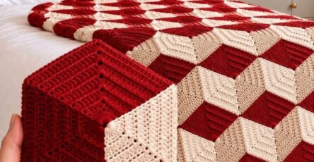 A geometric crochet blanket and matching cube pillow in red and cream, creating a 3D cube illusion, displayed on a bed with tassels on the blanket edge.