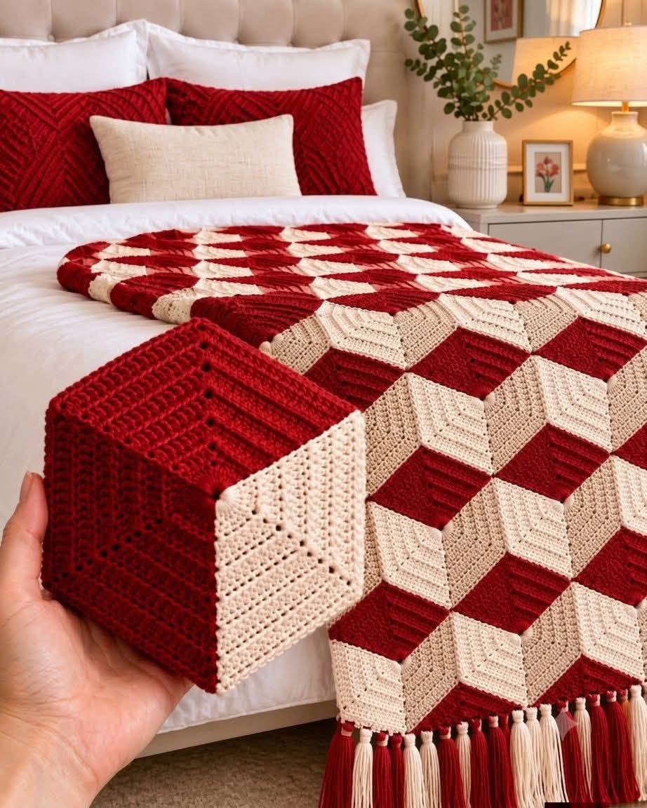 A geometric crochet blanket and matching cube pillow in red and cream, creating a 3D cube illusion, displayed on a bed with tassels on the blanket edge.