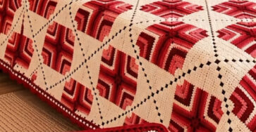 A hand holding a red and cream geometric granny square with a matching modular crochet blanket and pillows on a bed in the background.
