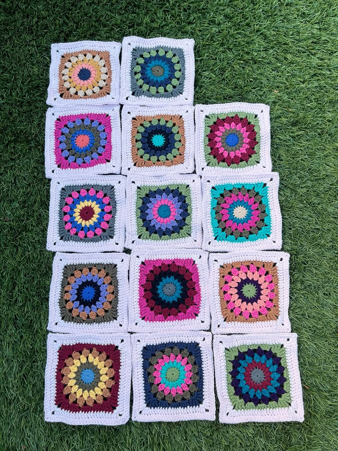 Fifteen crochet granny squares featuring colorful mandala circle centers and white borders, arranged on grass.