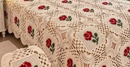 A crochet bedspread and pillow set featuring cream-colored granny squares with red rose motifs, draped neatly over a bed.