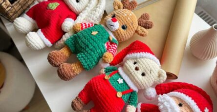Four crocheted Christmas plush toys including a bunny, reindeer, bear, and Santa Claus, all dressed in festive outfits, lying side by side on a table.