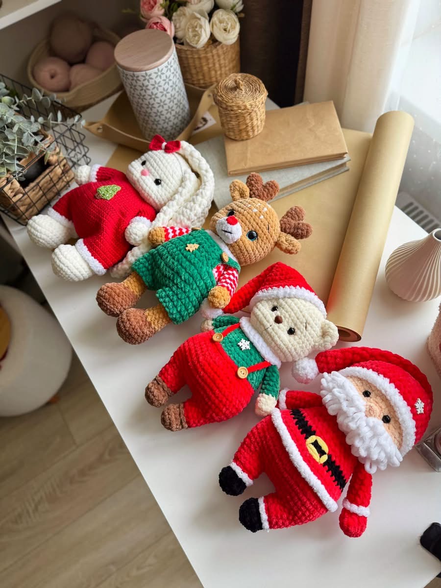 Four crocheted Christmas plush toys including a bunny, reindeer, bear, and Santa Claus, all dressed in festive outfits, lying side by side on a table.