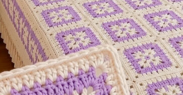 A lavender and cream granny square crochet bedspread with matching pillow covers, featuring floral-centered squares and a textured border.