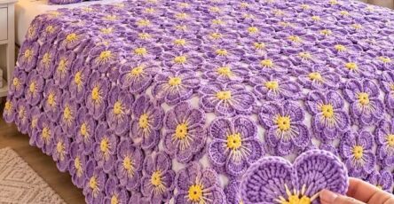 A hand holds a crocheted lavender and yellow flower motif in front of a bed covered with a matching crochet flower bedspread and pillows.