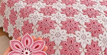A bedspread and pillows decorated with intricate pink and white crochet flower motifs; a hand holds a single crochet flower motif in the foreground.