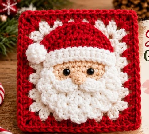 A crocheted granny square featuring a textured Santa Claus face with a fluffy white beard, red hat, and decorative border on a red background.