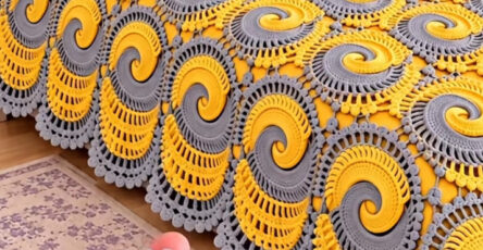 A hand holding a yellow and gray spiral crochet motif, shown as part of a bedspread on a neatly made bed with matching pillow.