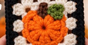 A crocheted granny square featuring an orange pumpkin with a green leaf and brown stem on a cream and black background.