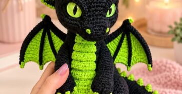 A crocheted dragon plush toy with black and bright green yarn, large green eyes, wings, belly scales, spikes, and textured details.