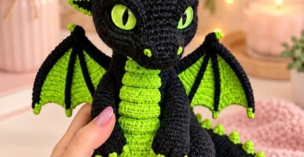 A crocheted dragon plush toy with black and bright green yarn, large green eyes, wings, belly scales, spikes, and textured details.