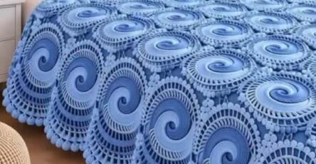 A luxurious blue crochet bedspread on a bed, featuring large swirling motifs, layered textures, and a scalloped border.