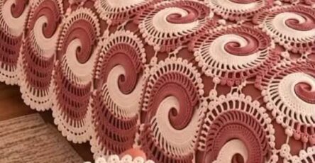 A bed covered in a handmade crochet bedspread featuring rose and cream swirling motifs, with a hand holding one motif in the foreground.