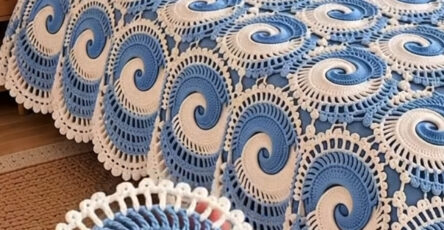 Hand holding a blue and white spiral crochet motif with a matching motif bedspread displayed on a bed in the background.