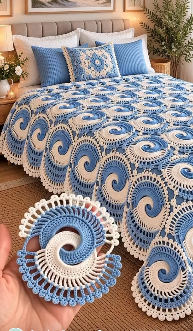 Hand holding a blue and white spiral crochet motif with a matching motif bedspread displayed on a bed in the background.