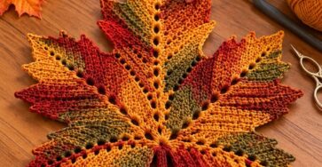 A large crocheted maple leaf in shades of orange, gold, green, and rust, laid flat on a wooden table as autumn decor.