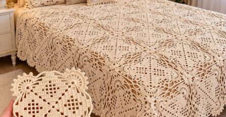 Hand holding a lacy crochet motif in front of a vintage-style crocheted bedspread made of joined square motifs in cream color.