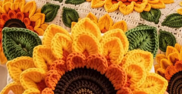 Hands holding a large 3D crocheted sunflower motif with a sunflower-themed blanket on a bed in the background.