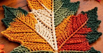 A large crocheted maple leaf placemat in orange, green, yellow, and cream, displayed as fall table decor with pumpkins and candles.