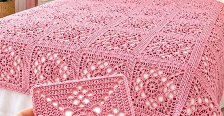 A hand holds a pink crocheted granny square in front of a pink crocheted bedspread and matching pillow covers on a bed.