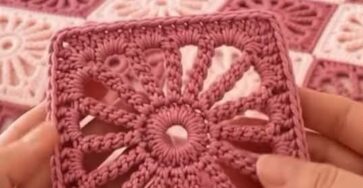 Two hands hold a sunburst motif crochet square in pink yarn with a matching pink and cream granny square blanket on a bed in the background.