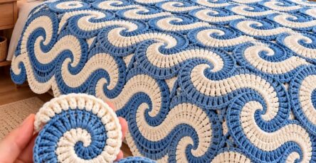 A hand holding a blue and cream crochet spiral motif in front of a bed covered with a matching crochet spiral blanket and pillows.
