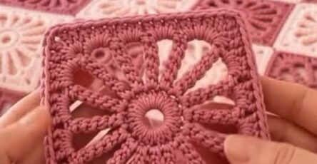 Two hands hold a sunburst motif crochet square in pink yarn with a matching pink and cream granny square blanket on a bed in the background.
