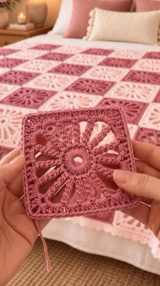 Two hands hold a sunburst motif crochet square in pink yarn with a matching pink and cream granny square blanket on a bed in the background.