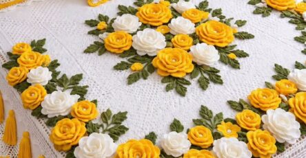 A crocheted bedspread and pillow set featuring large yellow and white 3D roses, green leaves, and a white lacy background with yellow tassels.