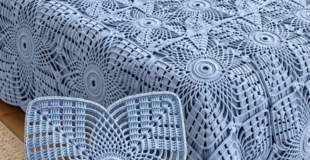 A hand holding a blue lacy square crochet motif, with a bedspread of similar motifs joined into a large blanket on a bed.