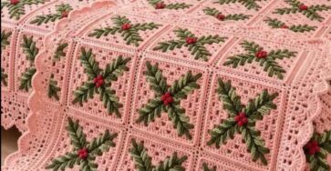 A cozy bed covered with a pink crochet blanket and pillows, each featuring green holly leaves and red berry motifs on granny squares, finished with a scalloped border.