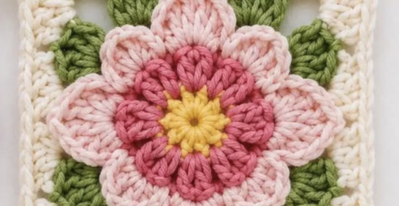 Crochet granny square with a layered pink and yellow flower and green leaves on a cream background.