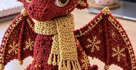A crocheted red dragon with gold horns, embroidered gold stars, large eyes, and a gold scarf, sitting on a desk.