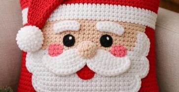 Crochet Santa Claus pillow with red hat, white beard, and cute facial features, displayed on a couch in a holiday setting.